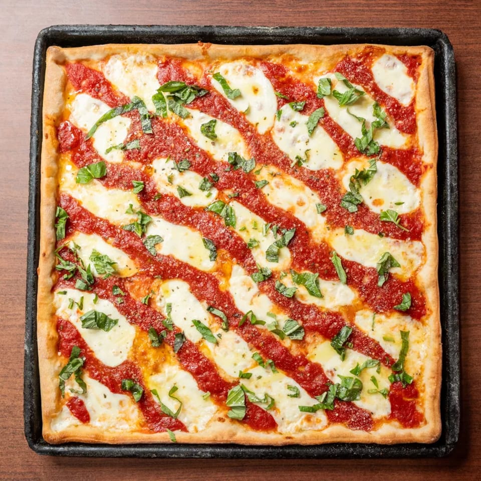 Square pizza with diagonal stripes of red sauce, white cheese, and fresh basil on a dark pan.