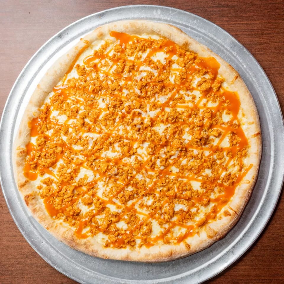 Round pizza with white cheese, orange crumbled chicken, and buffalo sauce drizzle on a metal pan.