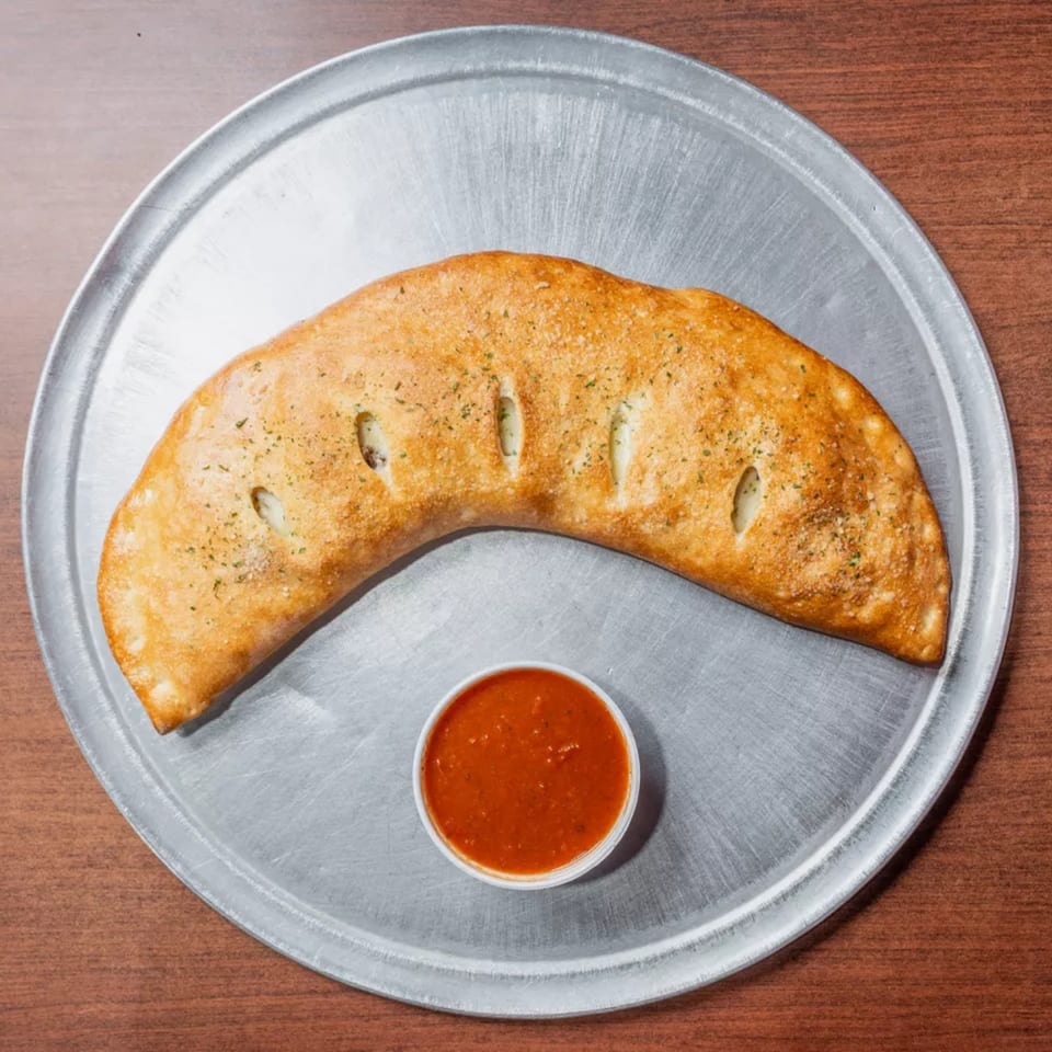 Golden calzone with herbs and vents, served with a cup of marinara sauce on a metal tray.
